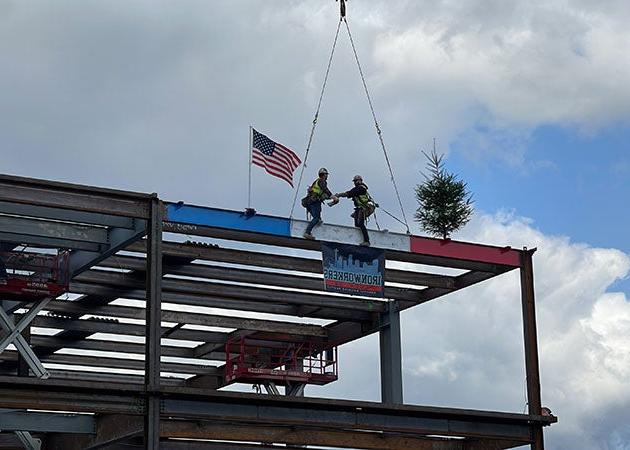 两个人站在钢架结构的一个角落的红、白、蓝三色梁上. 他们在握手. 一端是常青树，另一端是美国国旗.